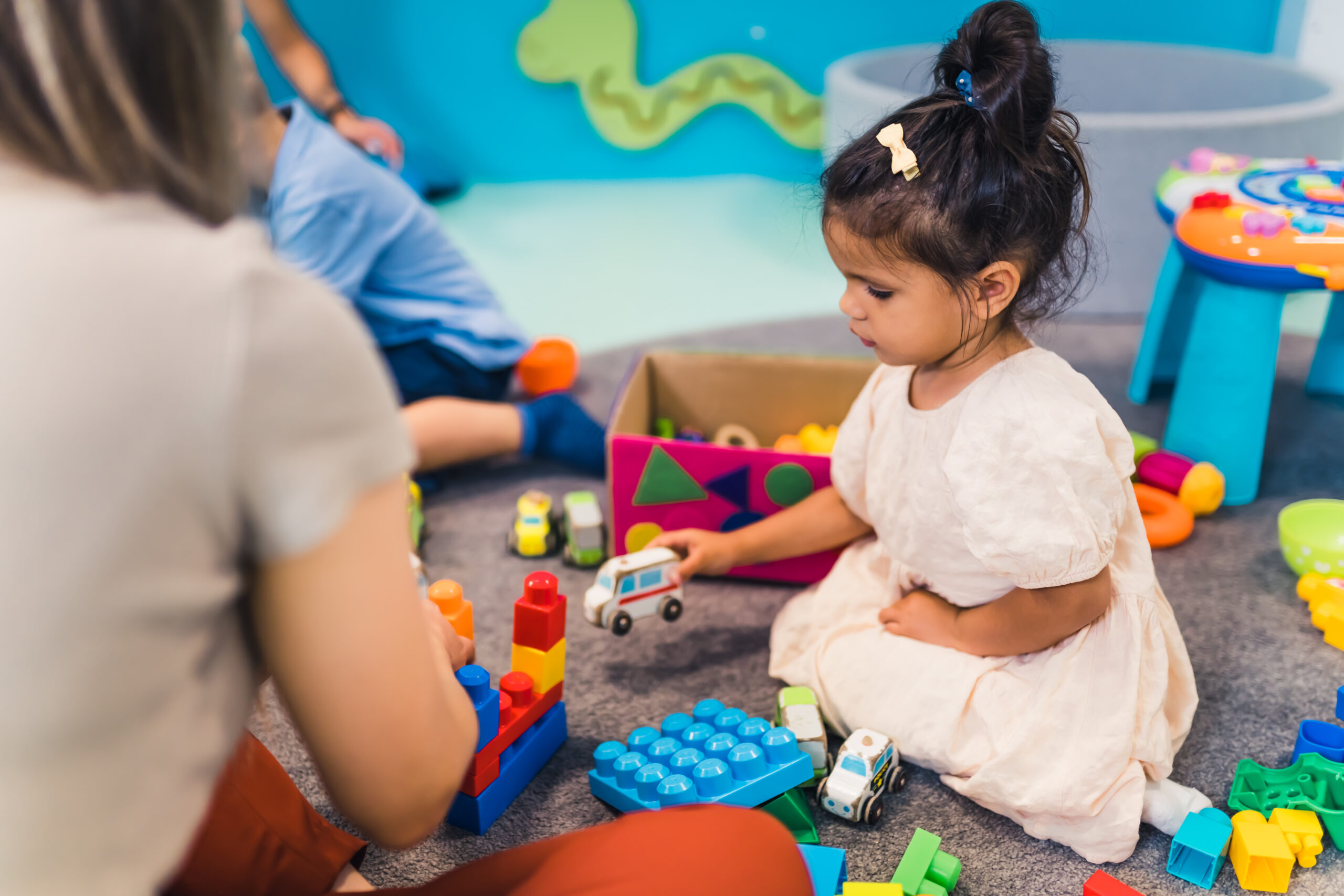 Learning through play at the nursery school. Toddler little girl and the teacher playing with colorful plastic playhouses, building blocks, cars and boats. Imagination, creativity, fine motor and gross motor skills development